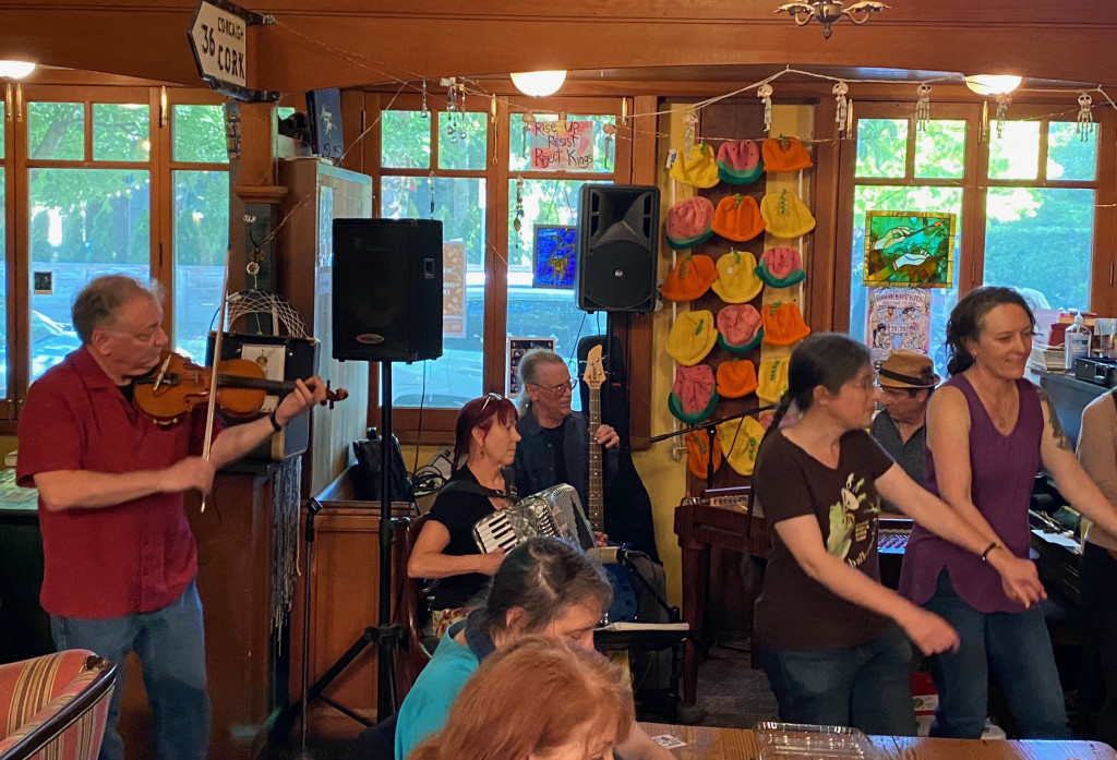 Trio Tsuica at Rose City Book Pub: standing at left, Brent Geary playing violin in a red short sleeved shirt; Kathy Fors on accordion in a black shirt, her glasses perched on her red hair; Peter Whitmore playing bass guitar in an upright position; Paul Beck in a porkpie hat behind two women doing a circle dance. The background is cluttered with PA speakers on stands, a display of knitted hats, windows showing glimpses of a summer afternoon street scene, and much other clutter typical of a neighborhood pub.